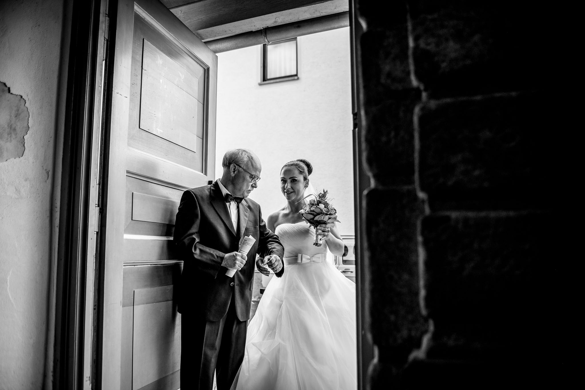 Eine Braut mit Blumenstrauß und eine ältere Frau stehen gemeinsam in einer offenen Tür, fotografiert in Schwarz-Weiß.